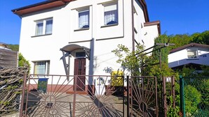 Ein zweigeschossiges Haus mit brauner Tür, einer kleinen Veranda und einem Metallzaun.