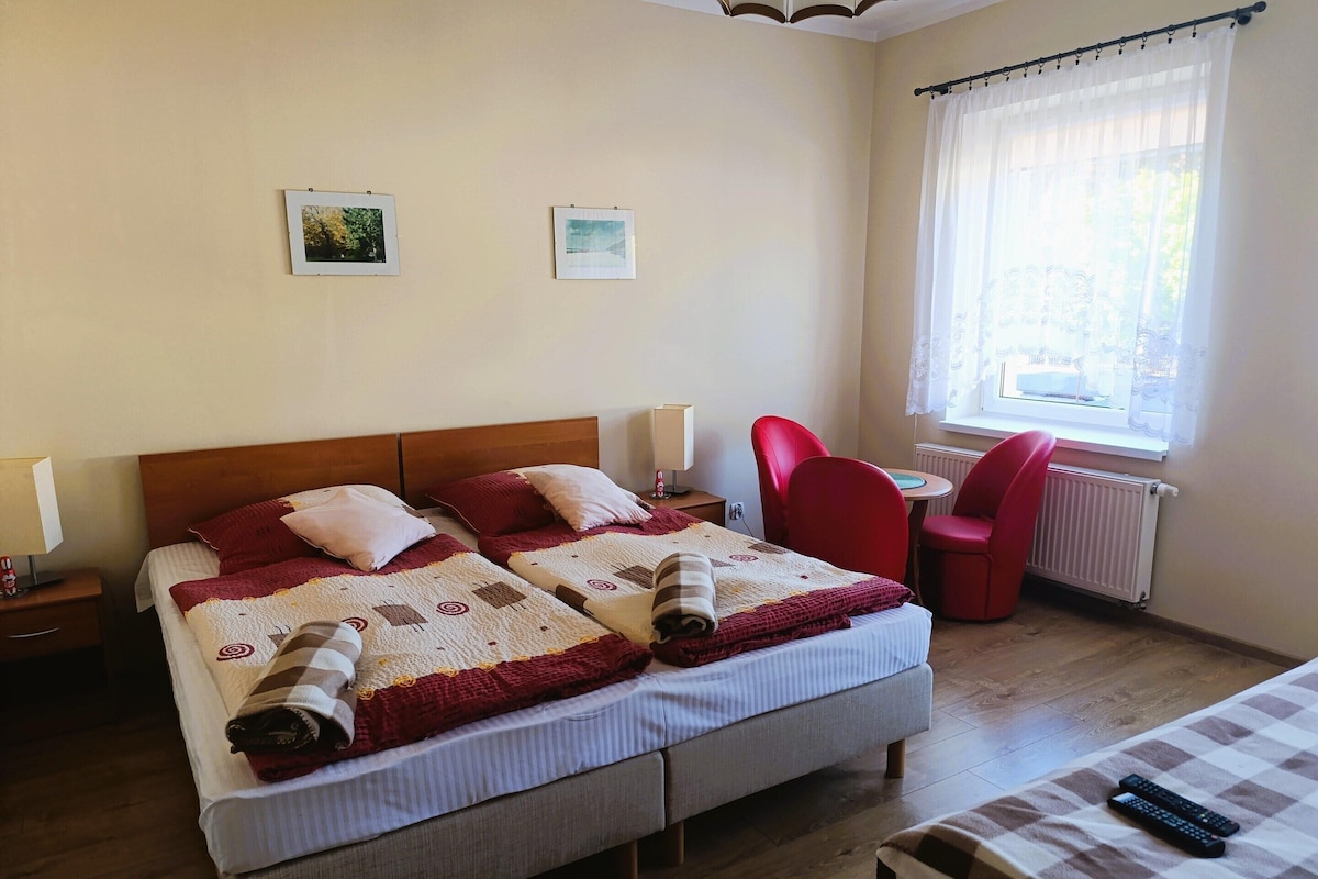 Ein Doppelbett mit Tagesdecke, zwei Nachttische mit Lampen, ein roter Sessel, ein kleiner Tisch mit Stuhl und ein Fenster mit Vorhängen.