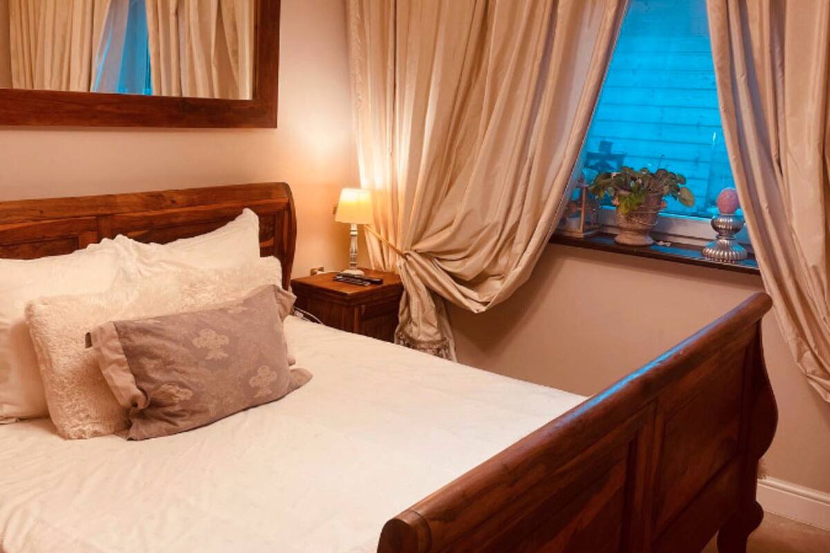 A neatly made bed with pillows, a bedside lamp, a window with curtains, and a potted plant on the windowsill.