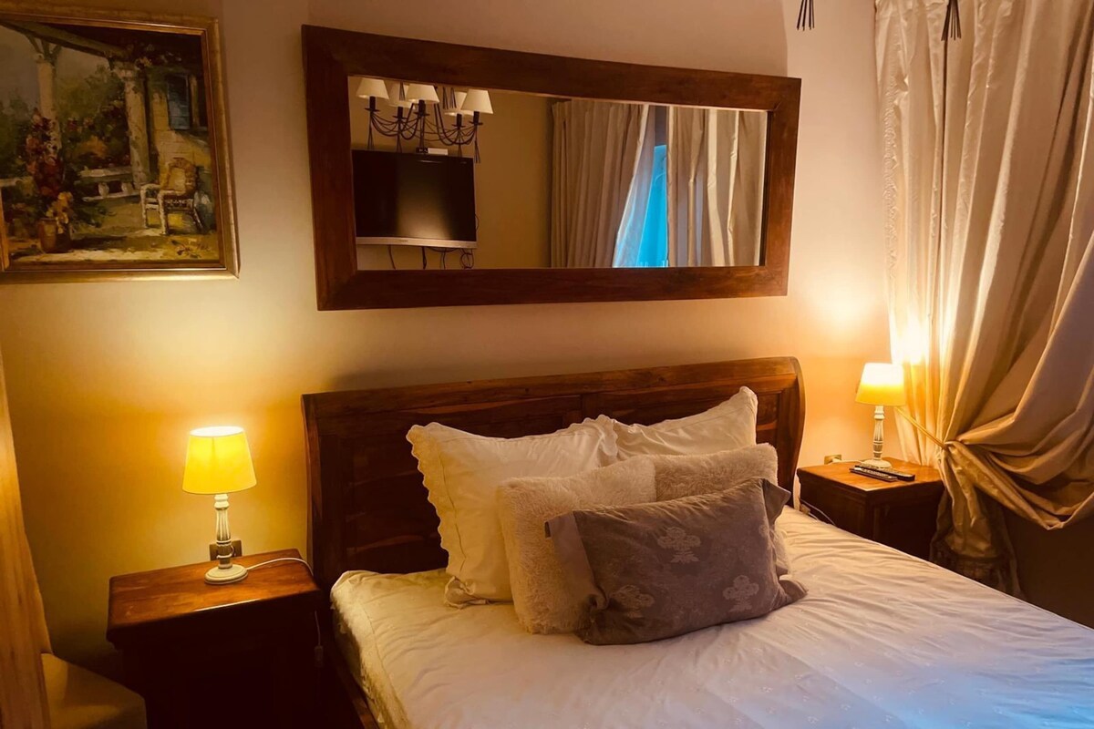 A neatly made bed with pillows, a bedside lamp, a nightstand, a painting on the wall, and a mirror above the bed.