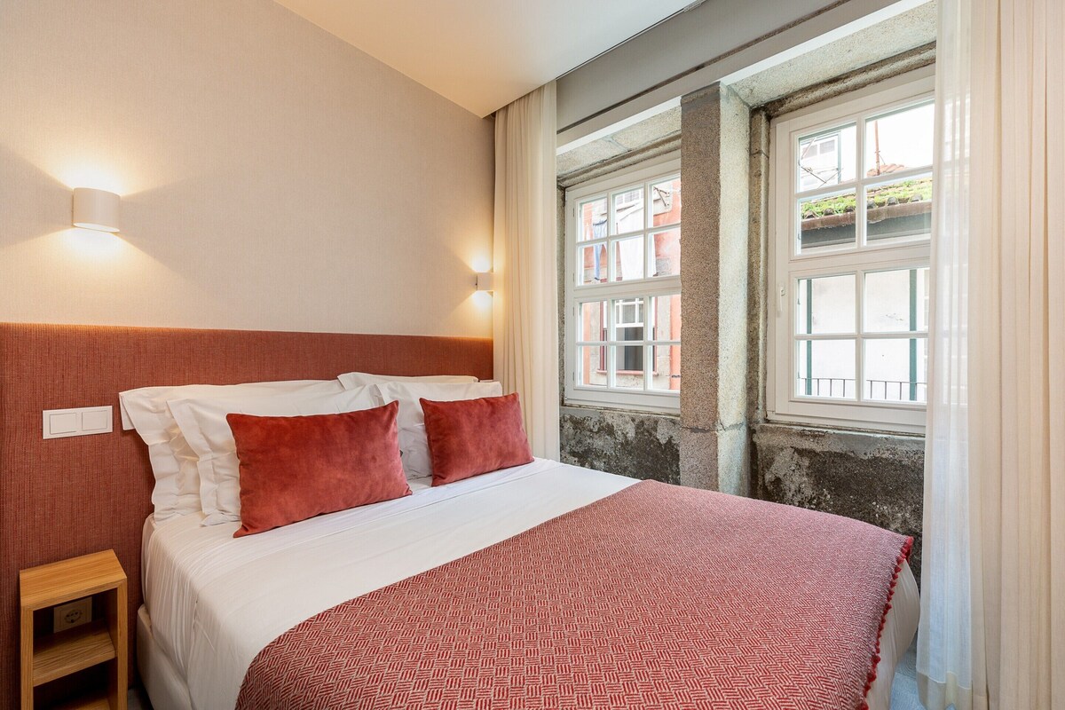 Ein ordentlich bezogenes Bett mit einem gemusterten Überwurf, zwei Wandleuchten, ein Nachttisch aus Holz und ein Fenster mit Blick auf Gebäude.