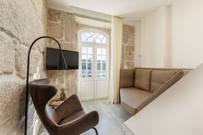 Quarto com parede de pedra, televisão montada, poltrona de couro marrom, sofá bege e porta branca com painéis de vidro.