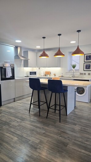 A modern kitchen with a central island, bar stools, pendant lighting, and a built-in washing machine. - Menai View Apartment (Y Felinheli)