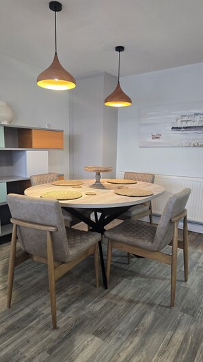 A dining area with a round table, six chairs, two pendant lights, and a wall-mounted radiator. - Menai View Apartment (Y Felinheli)