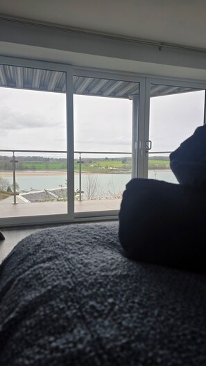 A room with a large window offering a view of a body of water and fields. - Menai View Apartment (Y Felinheli)