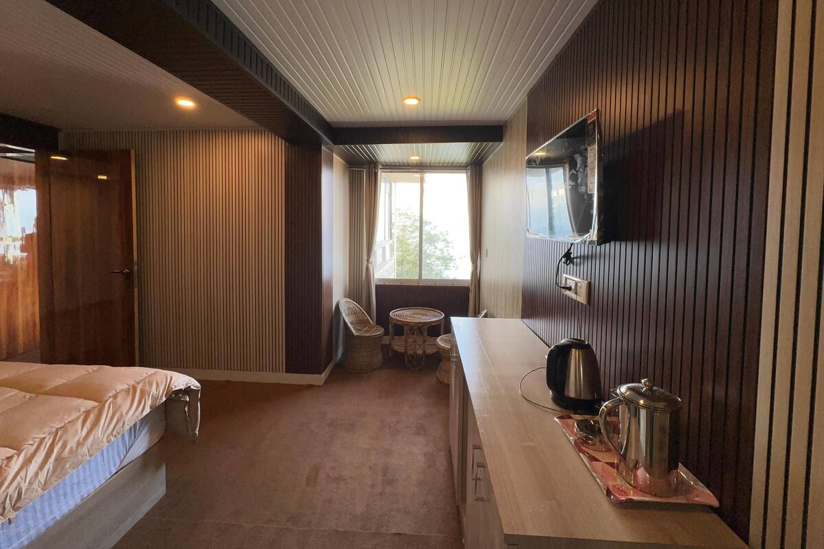 A bedroom with a bed, a TV mounted on the wall, a small table with a kettle, and a window with curtains.