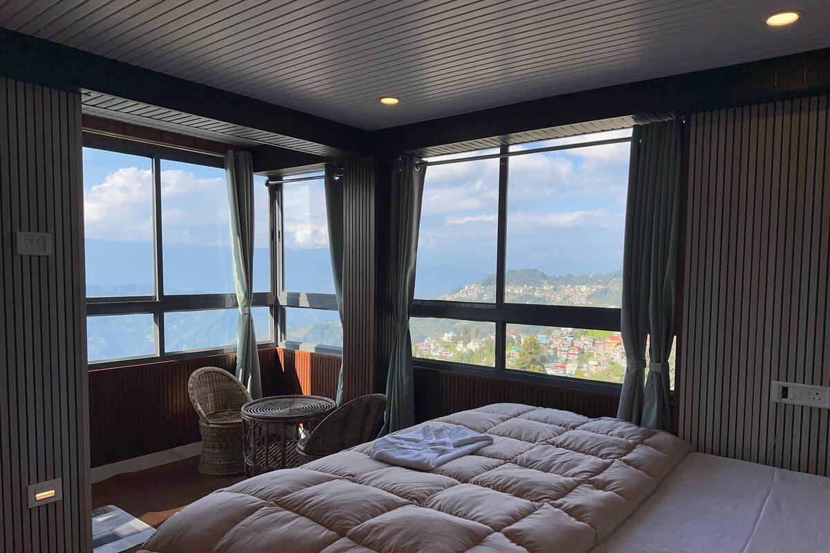 A bedroom with a large bed, a chair, and a view of a mountainous landscape through large windows.