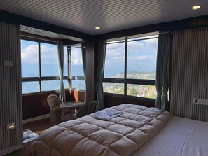 A bedroom with a large bed, a chair, and a view of a mountainous landscape through large windows. - Hotel Basera (Darjeeling)