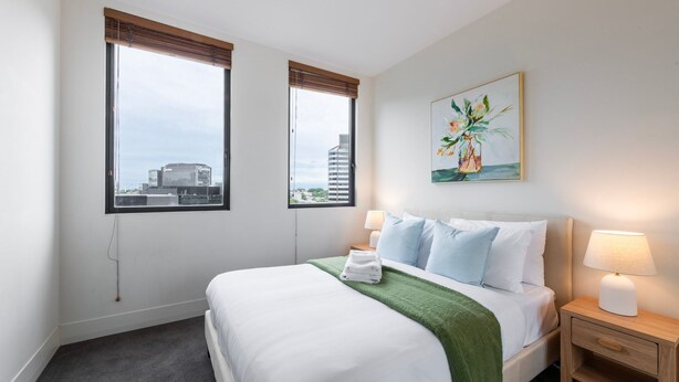 Habitación de hotel con una cama, dos ventanas con persianas, un cuadro en la pared y una mesita de noche con lámpara.