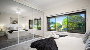 A bedroom with a large window, a bed, a nightstand, and a mirror. - Summer Wind - Beachside Stay Near Kendalls Beach (Kiama)