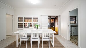 A dining area with a white table and chairs, a potted plant, and a view of a kitchen through a glass door. - Summer Wind - Beachside Stay Near Kendalls Beach (Kiama)