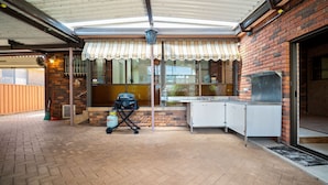 A covered patio area with a grill, a water heater, and a brick wall. - Summer Wind - Beachside Stay Near Kendalls Beach (Kiama)