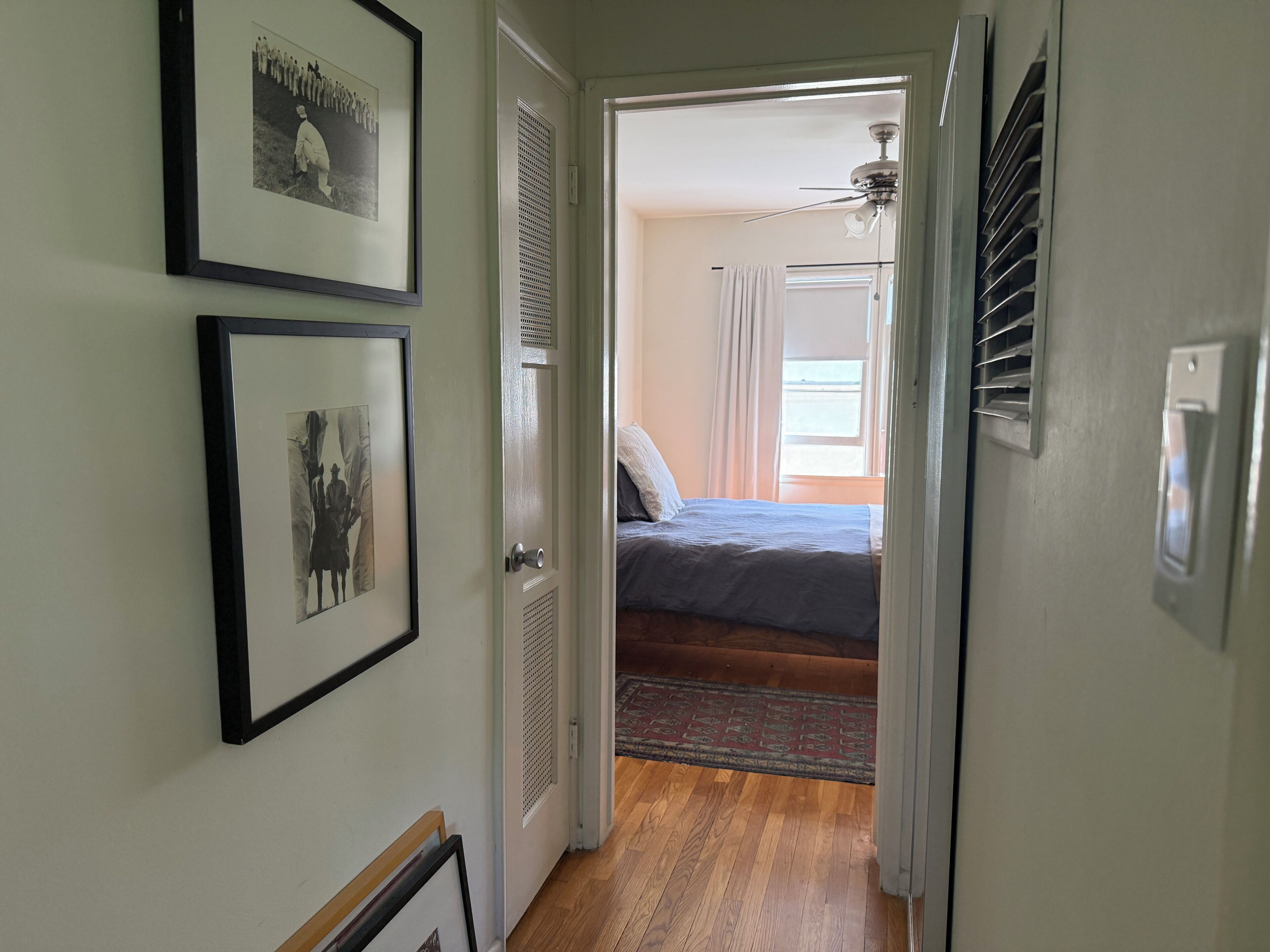 A hallway leading to a bedroom with a bed, a ceiling fan, and a window with curtains.