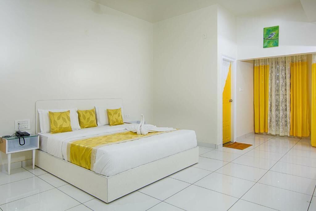 Una habitación de hotel con una cama blanca, almohadas amarillas, una mesita de noche blanca y un cuadro verde en la pared.
