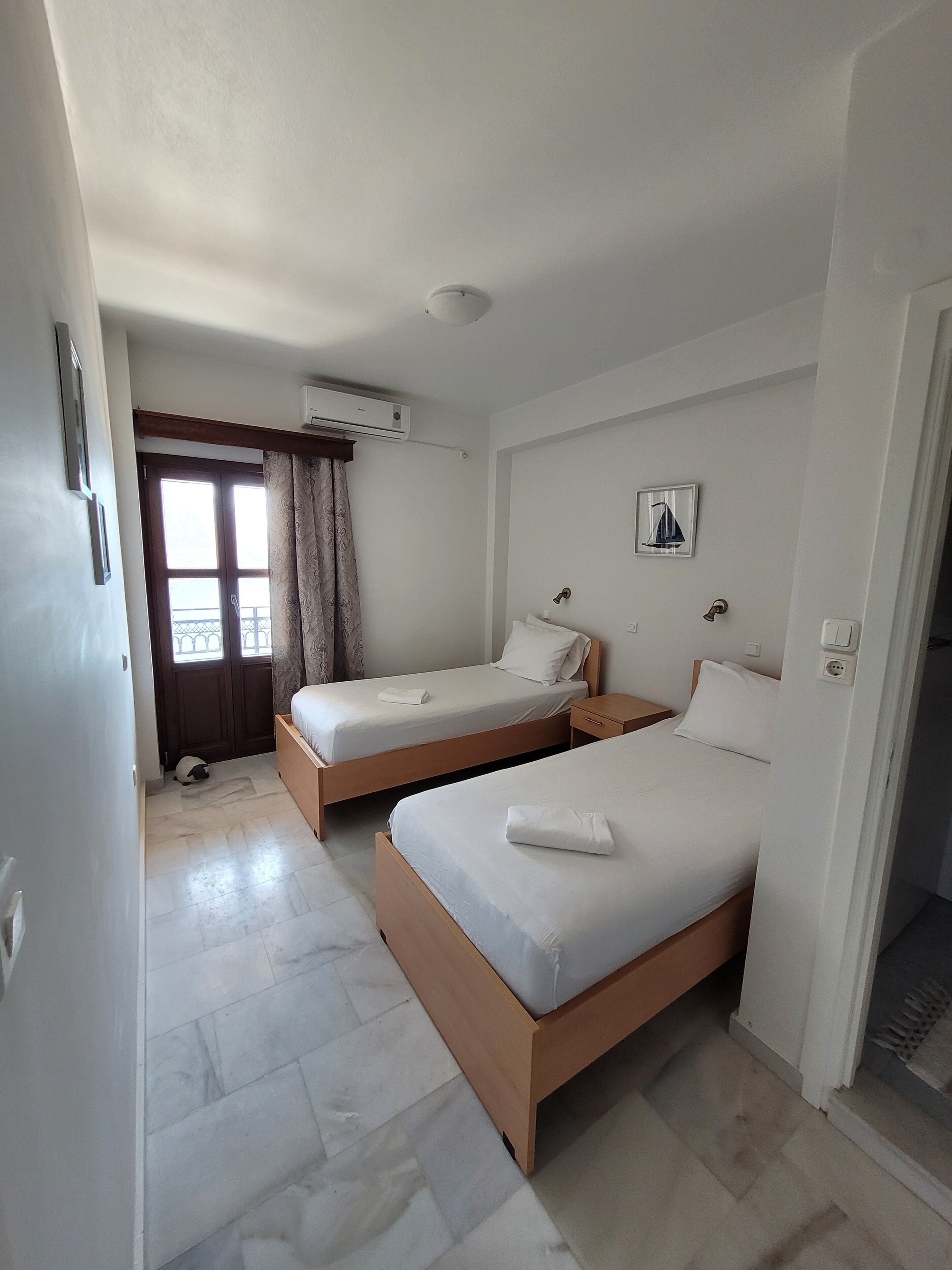 A hotel room with two single beds, a wooden floor, a ceiling fan, an air conditioning unit, a window with curtains, and a small bedside table with a lamp.