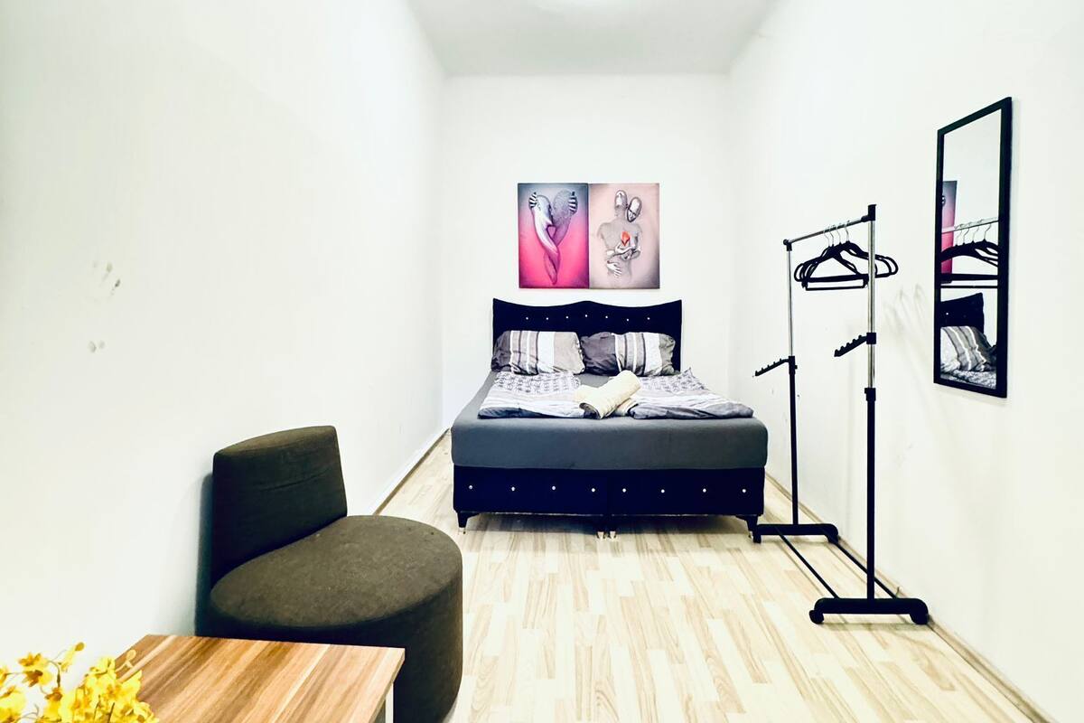 A small, modern room with a bed, a chair, a table with flowers, and a mirror on the wall.