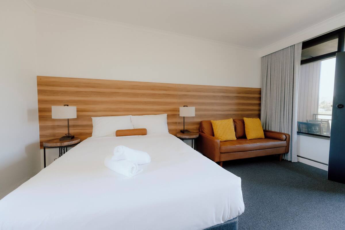 Une chambre d’hôtel avec un grand lit, deux tables de chevet avec des lampes, un canapé en cuir marron et une fenêtre avec des rideaux.