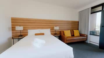 Een hotelkamer met een groot bed, twee nachtkastjes met lampen, een bruine leren bank en een raam met gordijnen.