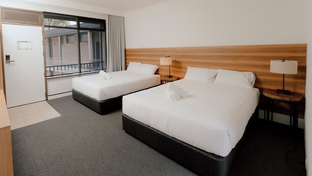 A hotel room with two beds, a wooden headboard, a small table with a lamp, and a window with curtains. - Canberra Park Motel (Watson)