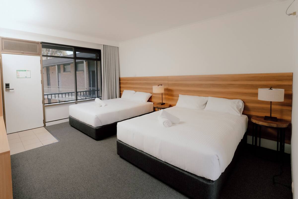 Une chambre d’hôtel avec deux lits, une tête de lit en bois, une petite table avec une lampe et une fenêtre avec des rideaux.