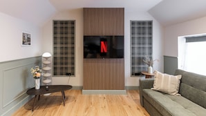 A modern living room with a flat-screen TV mounted on a wooden panel, a grey sofa, a round coffee table, and a vase with flowers. - New Apartment in Guildford (Guildford)