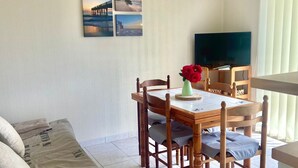 A living room with a sofa, a dining table with chairs, a ceiling fan, an air conditioning unit, a TV, and a wall with framed pictures.