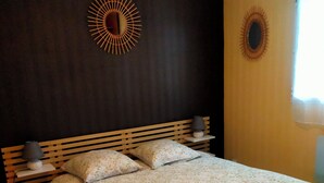 A bedroom with a wooden bed frame, a large floral bedspread, two bedside lamps, and a sunburst mirror on the dark wall.
