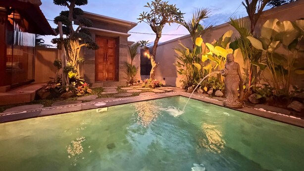 A swimming pool with a stone border, surrounded by tropical plants and a wooden structure. - Villa Uma Asri (Ubud)