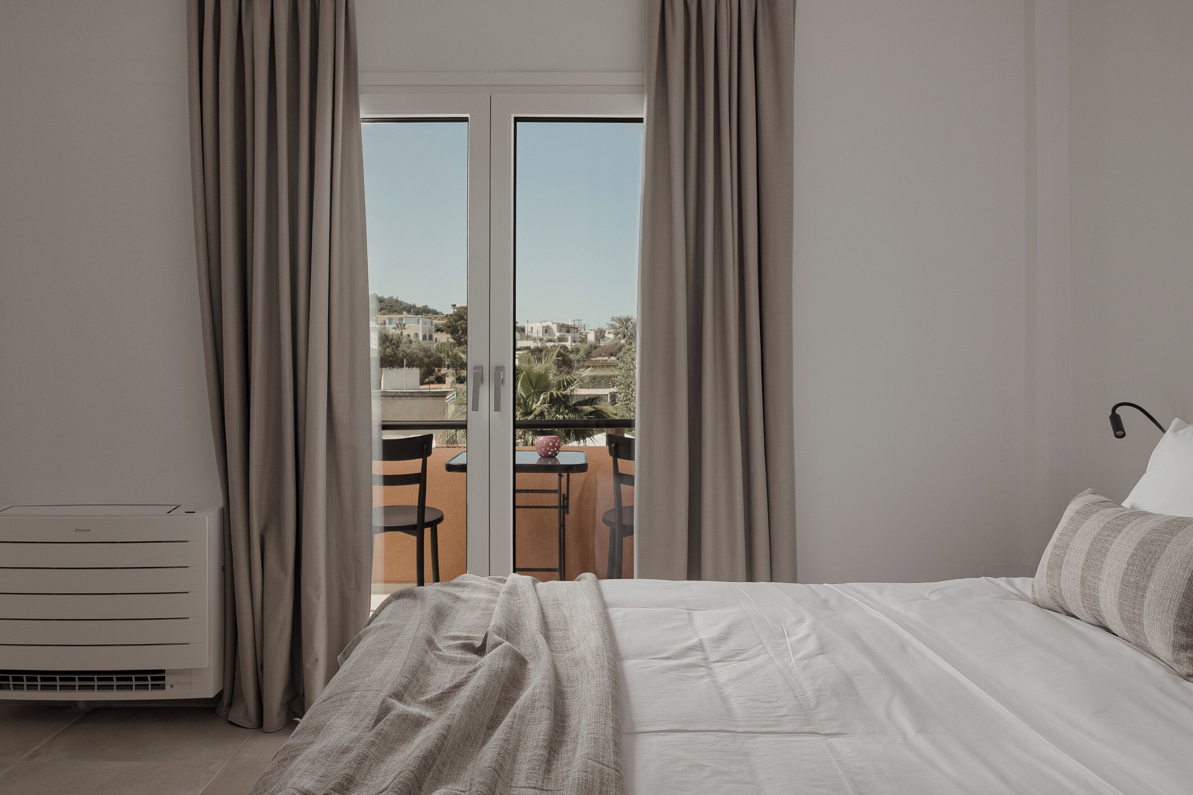 A hotel room with a bed, a radiator, and a view of the city through large windows.