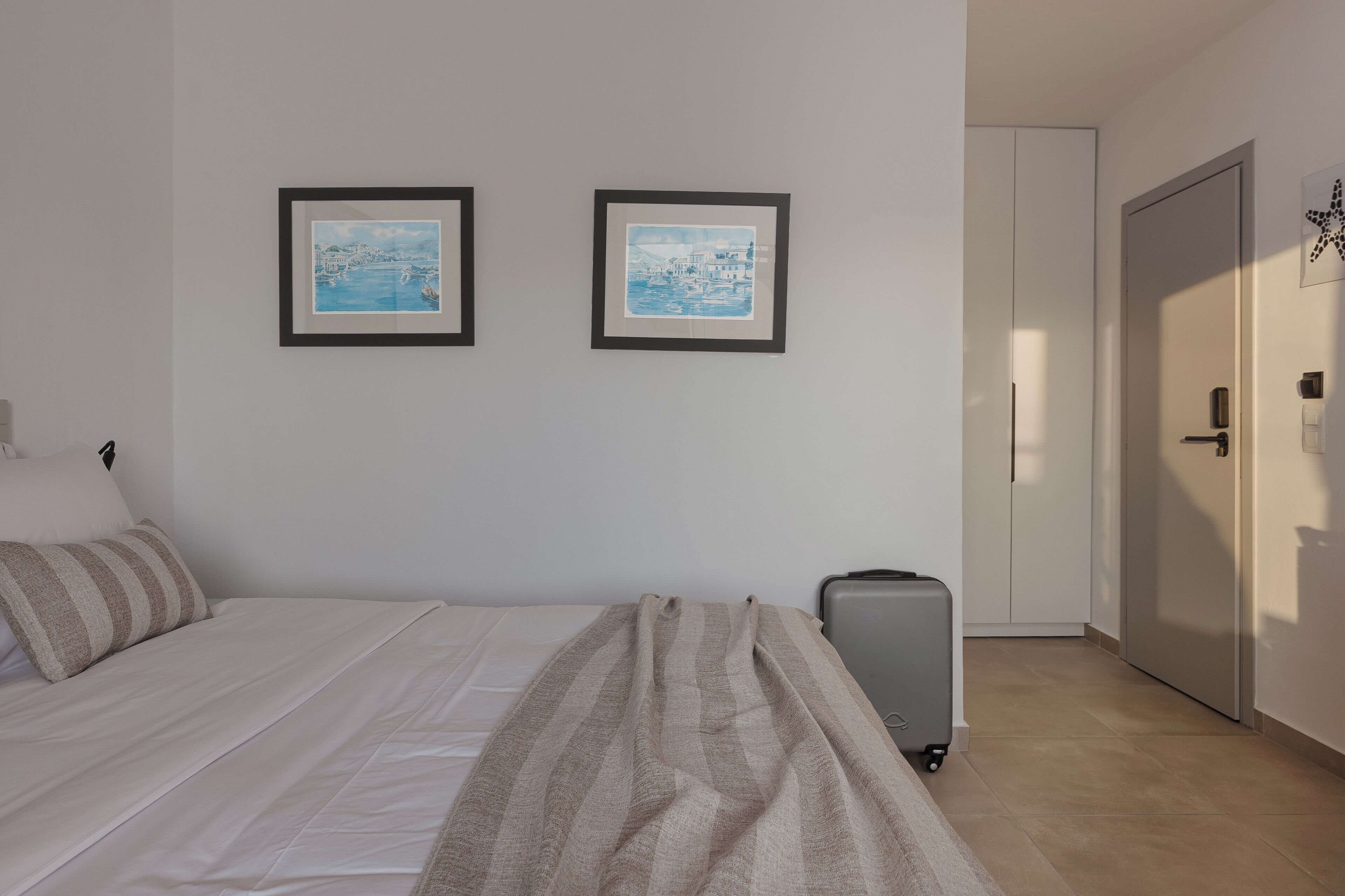 Ein Hotelzimmer mit einem Bett, einem Koffer, zwei gerahmten Bildern an der Wand und einer Tür.