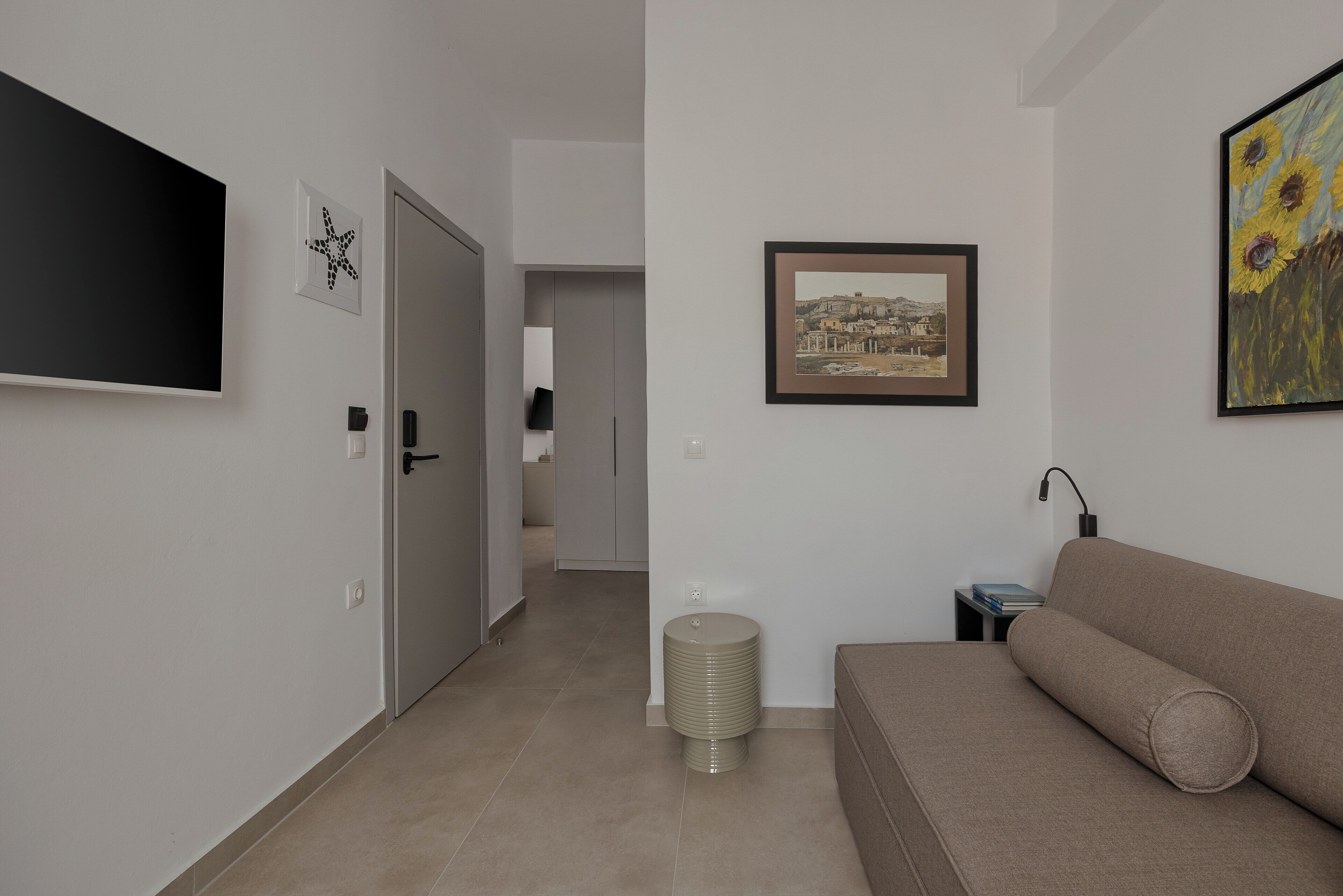 Un salon moderne avec un canapé, un téléviseur à écran plat, un tableau encadré représentant un paysage urbain et une petite table avec une lampe.