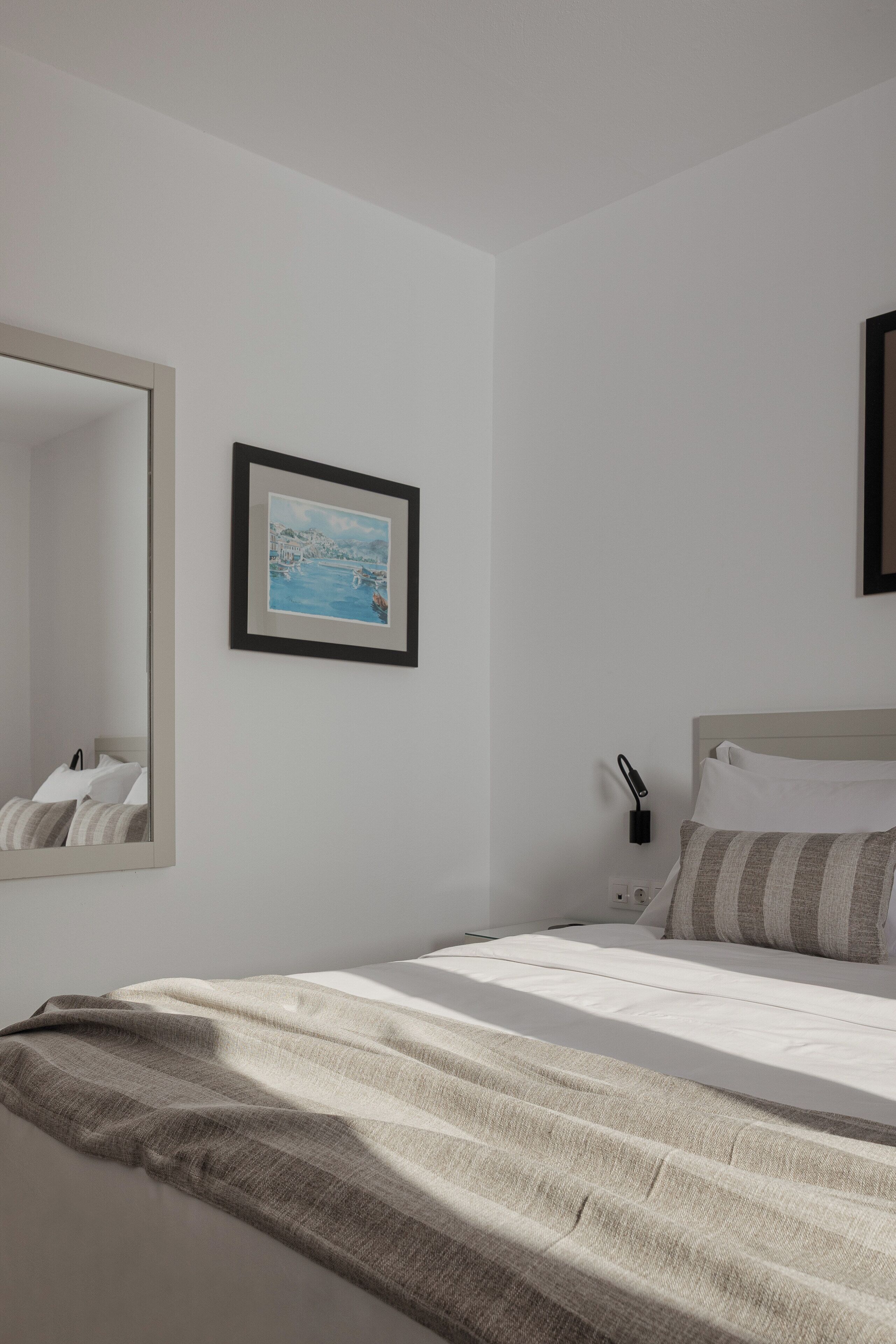 A bedroom with a bed, a bedside table, a nightstand, a wall-mounted lamp, a framed picture, and a mirror.