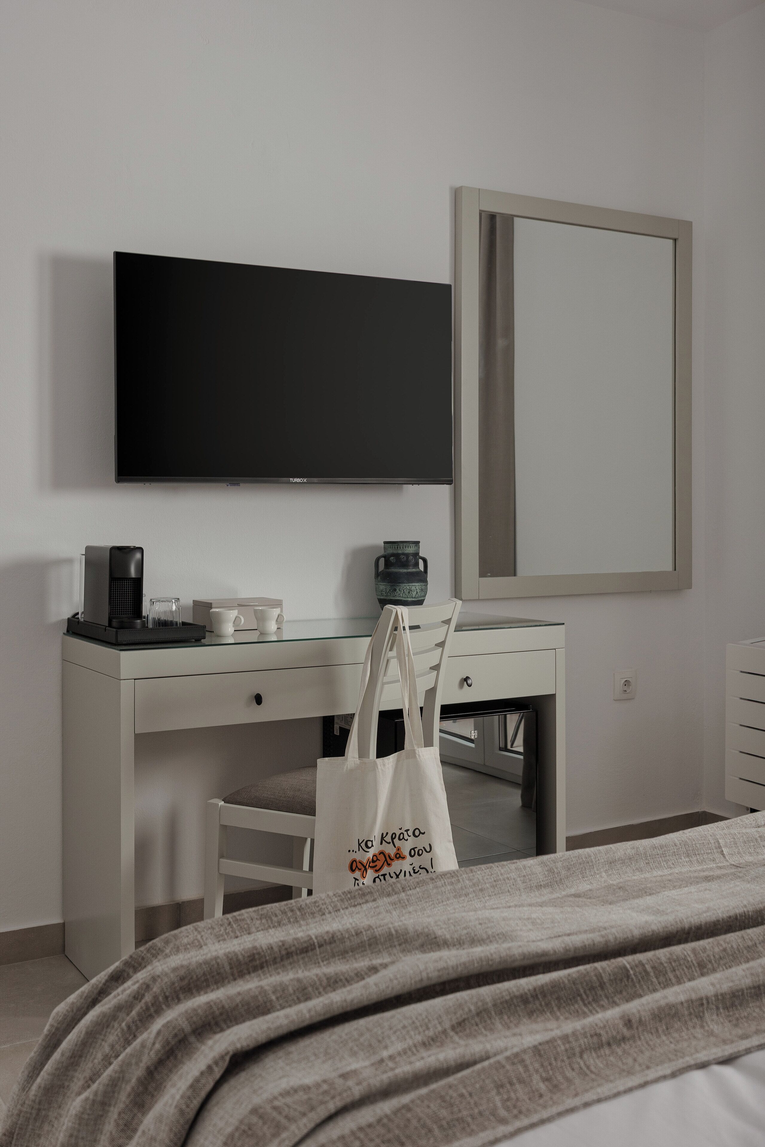 Une chambre d’hôtel comprenant un lit, un bureau avec une machine à café, une télévision, un miroir et un sac en toile blanc.