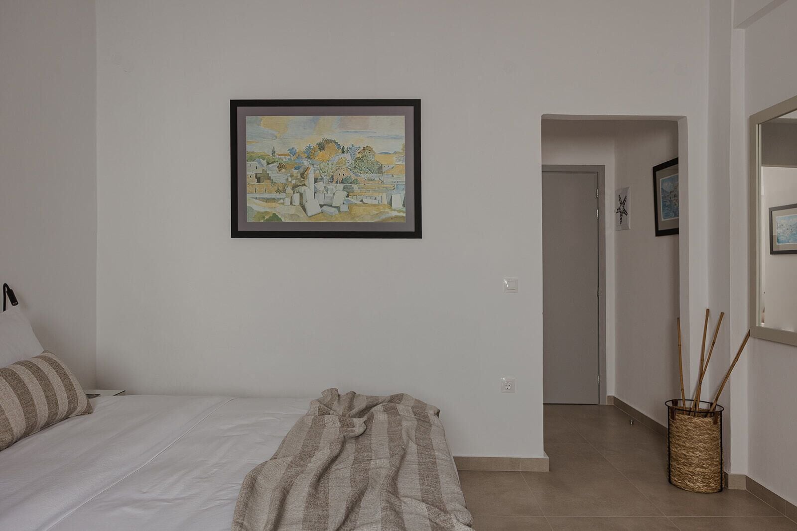 A bedroom with a bed, a blanket, a framed painting, a door, and a wicker basket.