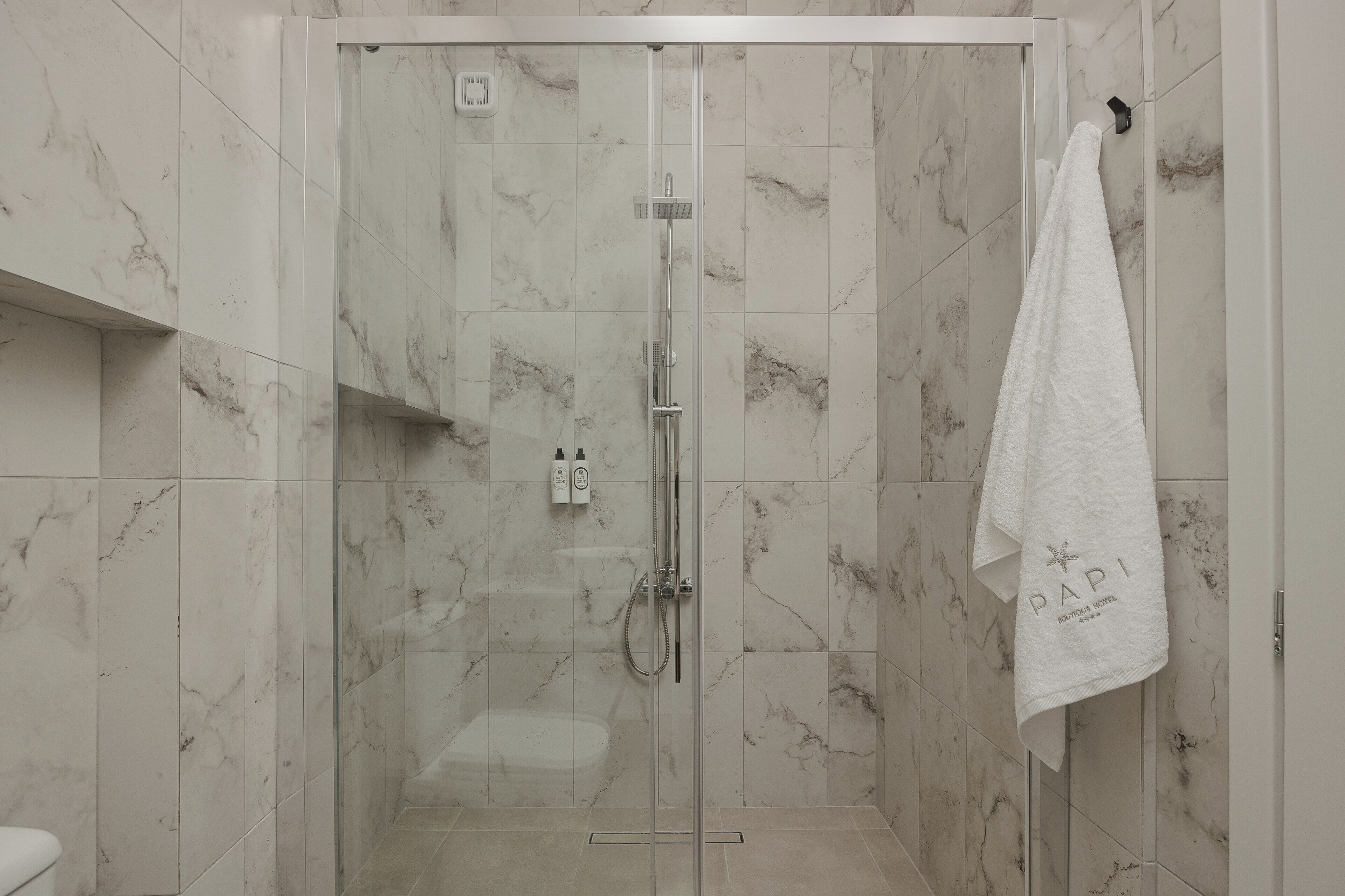 Ducha envuelta en vidrio con cabezal de ducha manual, una toalla colgada en un gancho y pared con azulejos de mármol.