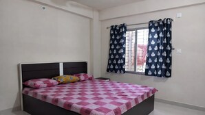 A bedroom with a bed, a window with patterned curtains, and a tiled floor. - Arjun Guest House (Bidhannagar)