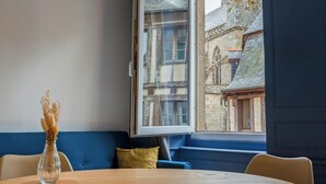 Una habitación con una mesa redonda de madera, un sofá azul, un florero transparente con plantas secas y una ventana con vista a un edificio histórico.