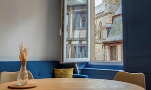 Una habitación con una mesa redonda de madera, un sofá azul, un florero transparente con plantas secas y una ventana con vista a un edificio histórico.
