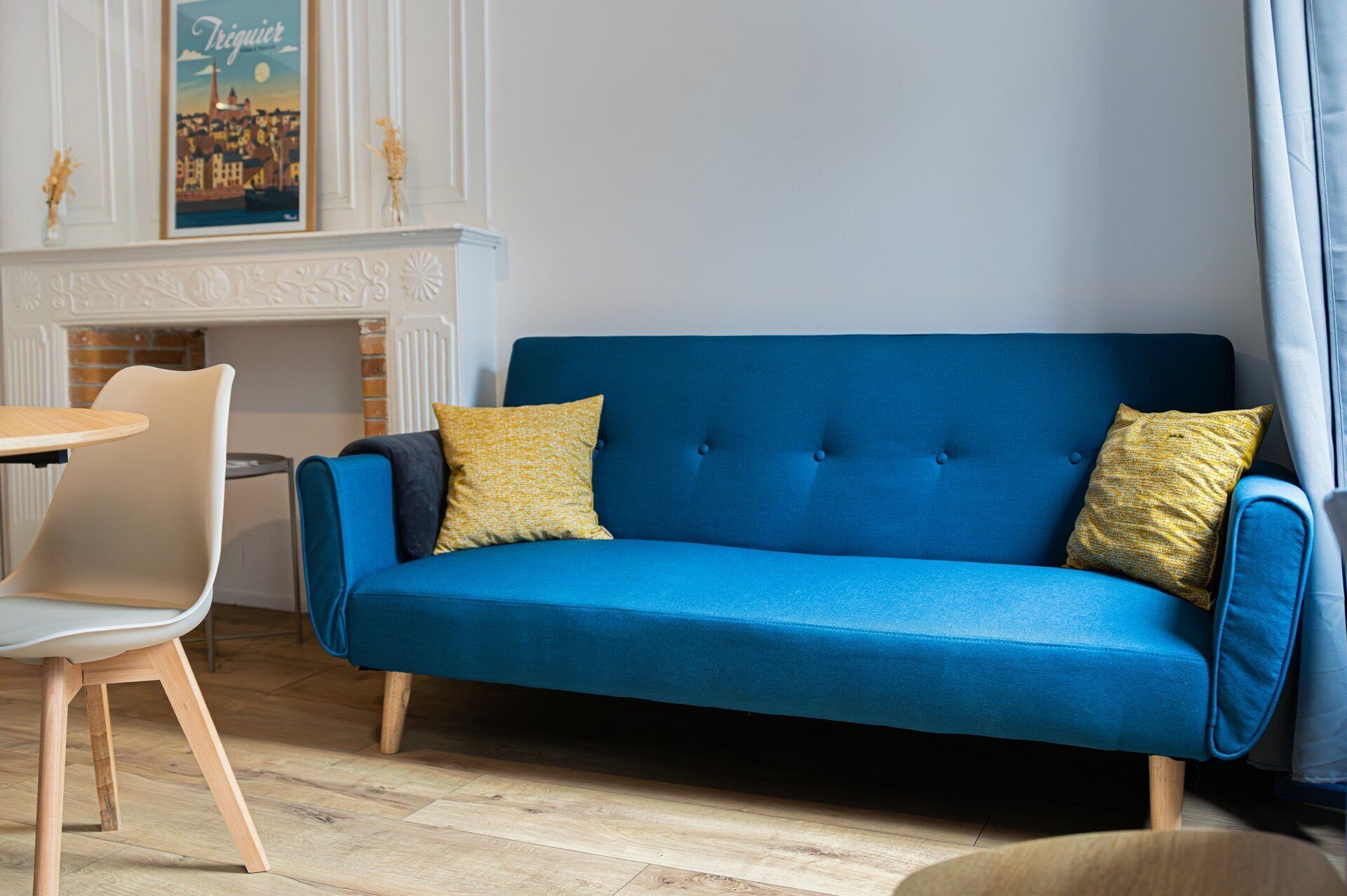 A blue sofa with two yellow pillows, a white chair, and a small round table in a room with a fireplace and a poster on the wall.