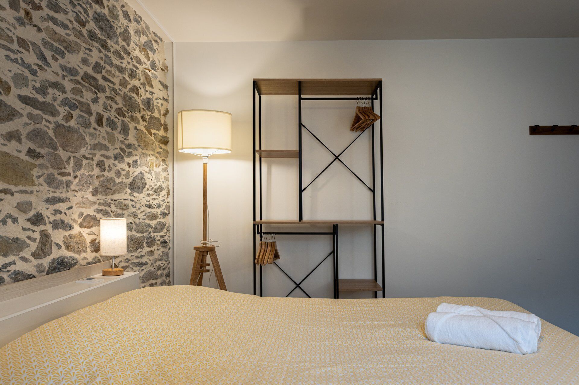 Una camera da letto con parete in pietra, un letto con copriletto decorato, una lampada da terra e un mobiletto a mensole in legno con abiti.