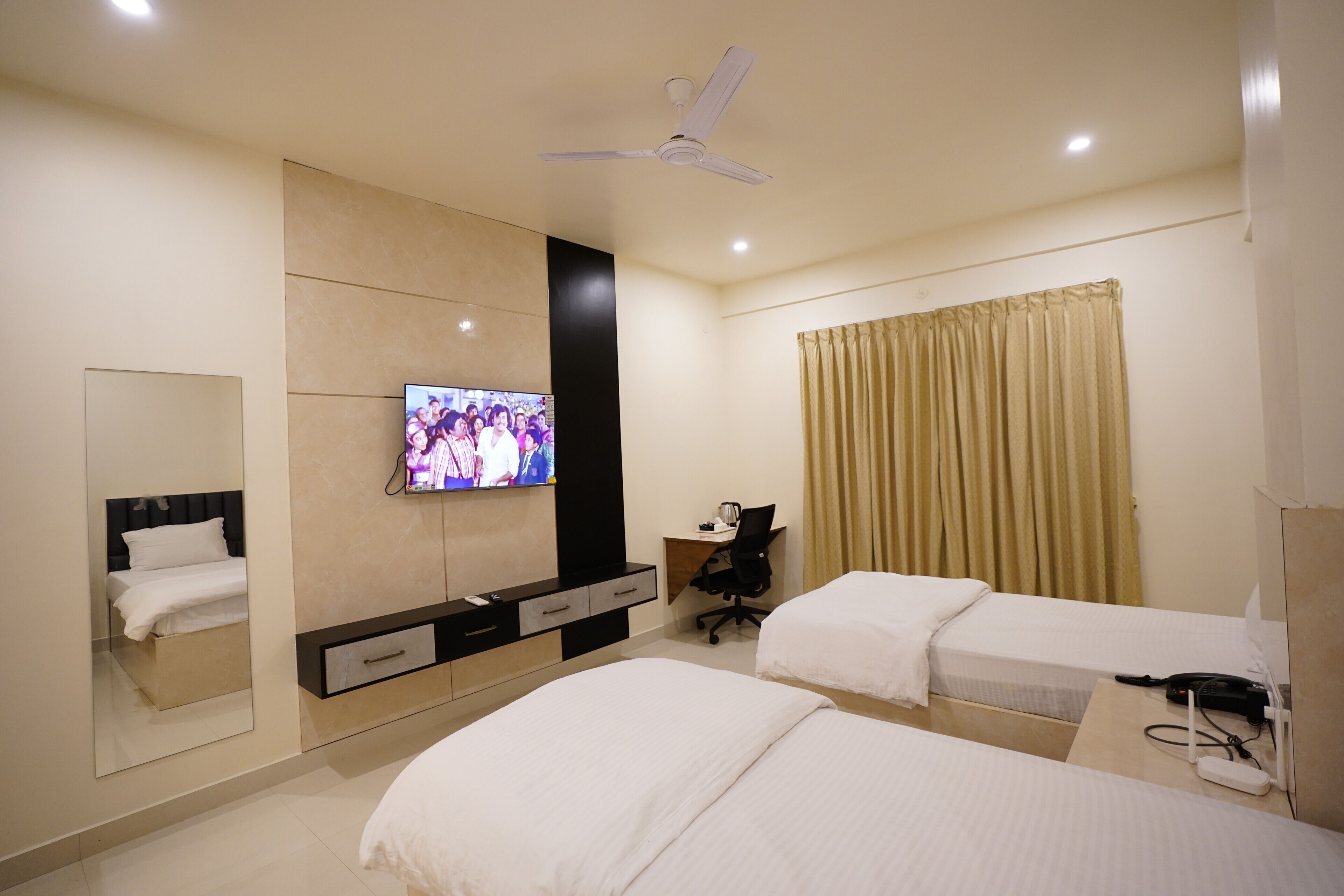 A hotel room with two beds, a desk, a television, a ceiling fan, and a window with curtains.
