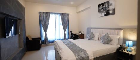 A modern hotel room with a bed, a desk, a chair, a TV, a ceiling fan, and a painting on the wall.