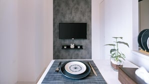 Un comedor moderno con un televisor de pantalla plana montado en una pared texturizada, un estante pequeño con un reloj y una planta, y una mesa de comedor dispuesta con platos y cubiertos.