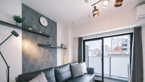 Una sala de estar moderna con un sofá de cuero negro, un reloj de pared, estanterías con plantas y un amplio ventanal con vistas a la ciudad.