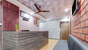 Ein Empfangsbereich mit Holzbude, Deckenventilator, Empfangsschild, Wand-Fernseher und einer Sitzecke mit gepolsterten Bänken.
