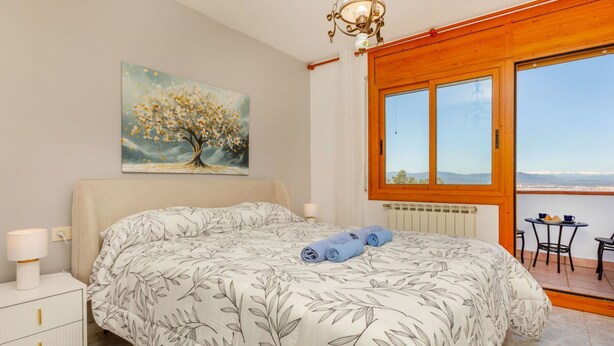Un dormitorio con cama, mesita de noche, lámpara, un cuadro y vistas al exterior.
