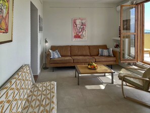 Ein Wohnzimmer mit einem braunen Sofa, einer gemusterten Bank, einem Couchtisch mit Obst und einem Fenster mit Aussicht.