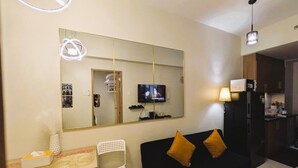 Un espace de vie compact doté d’un mur miroir, d’un téléviseur, d’un canapé noir avec deux coussins, d’une petite table blanche et d’une chaise.