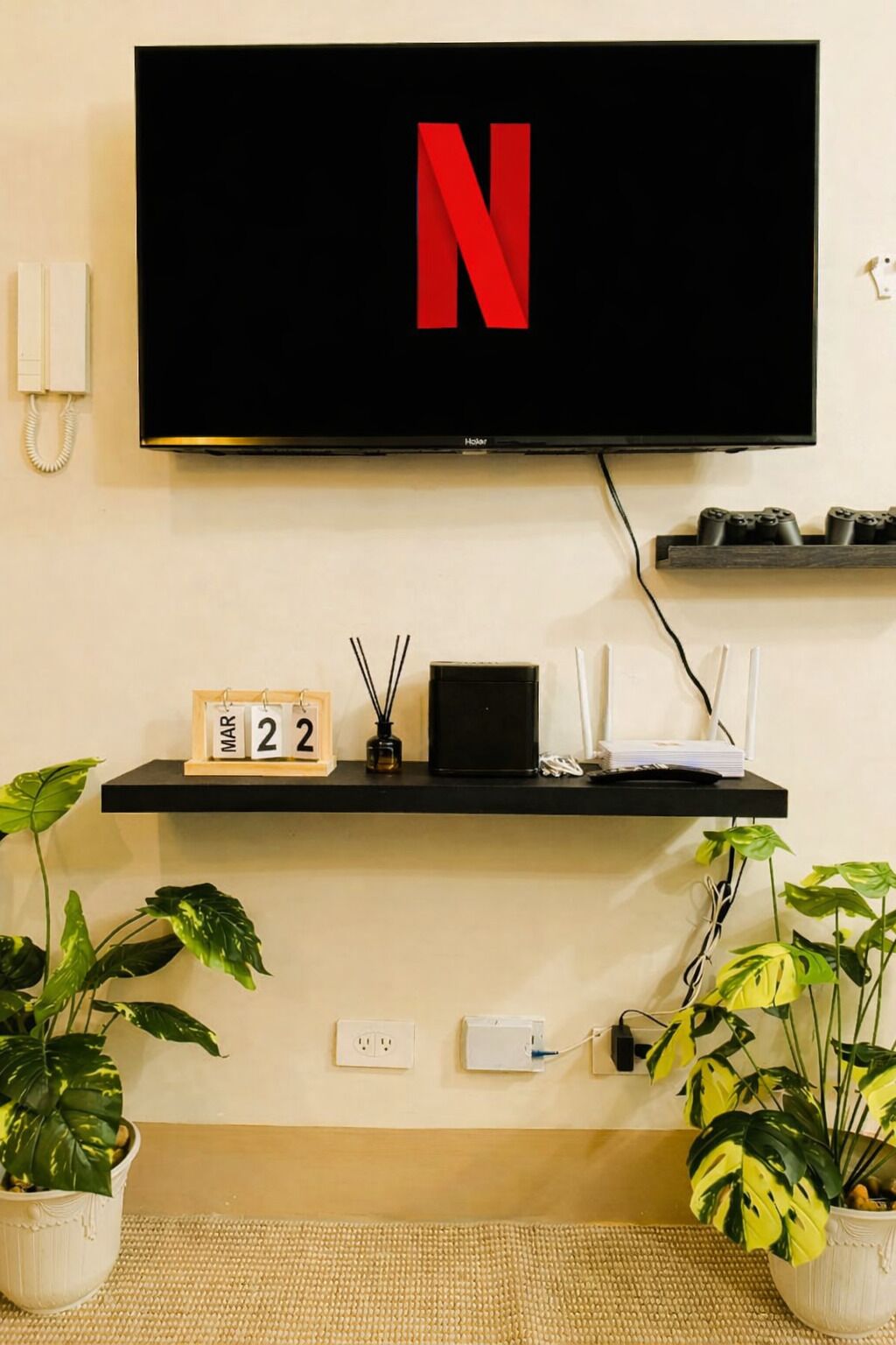 A flat-screen TV mounted on a wall with a shelf below displaying a calendar, a small speaker, and a plant.