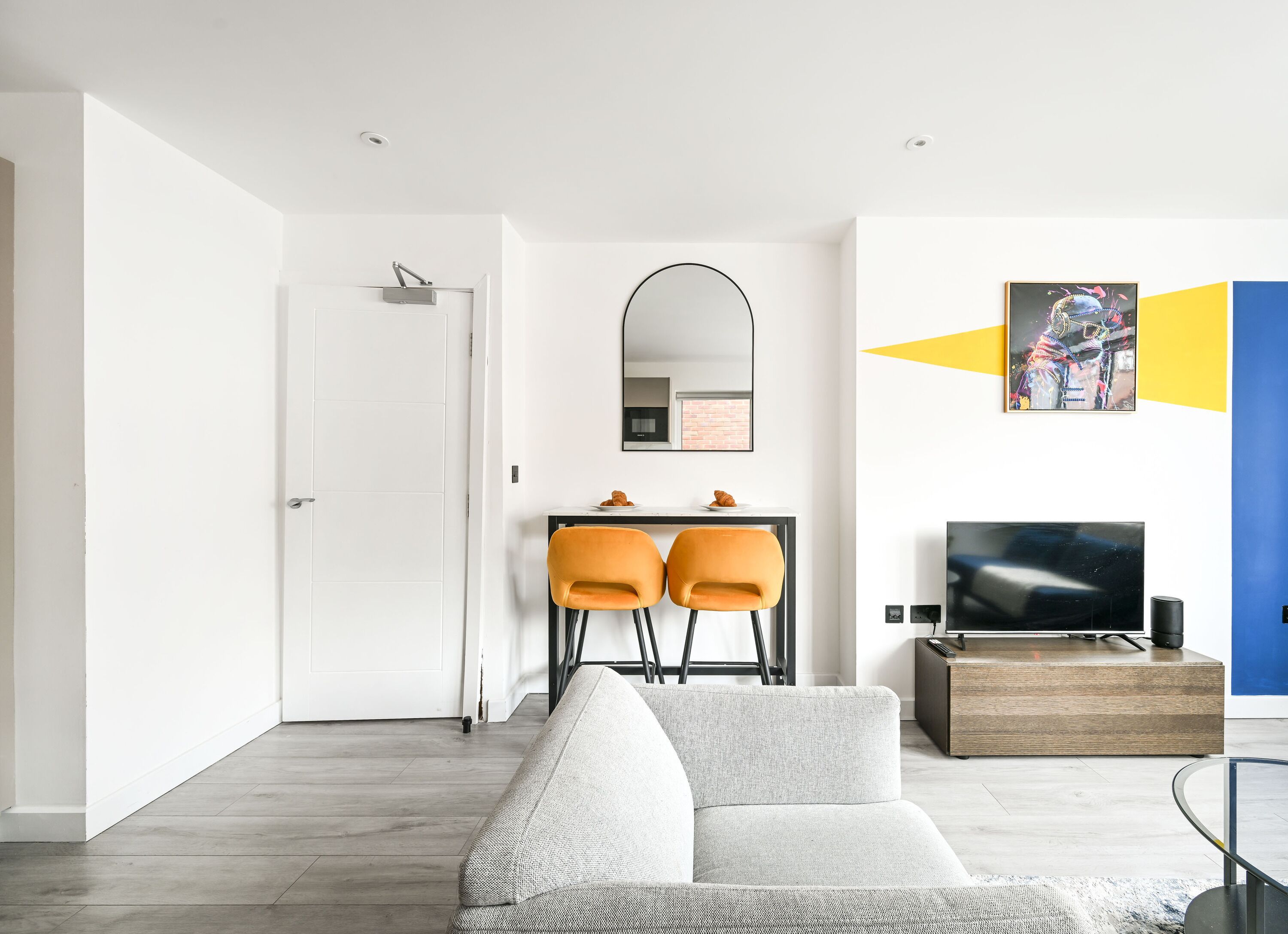 Een moderne woonkamer met een bank, een eethoek met twee stoelen, een televisie en een spiegel aan de muur.